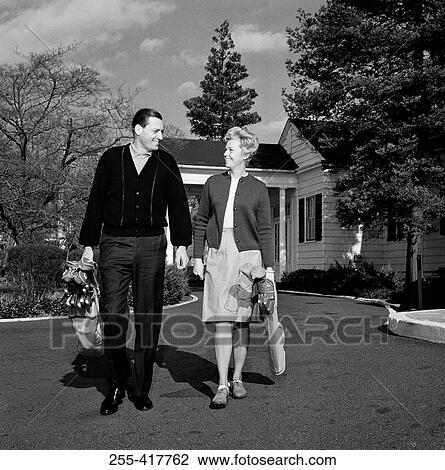 Stock Image - Mid adult couple walking side by side, carrying golf clubs. Fotosearch