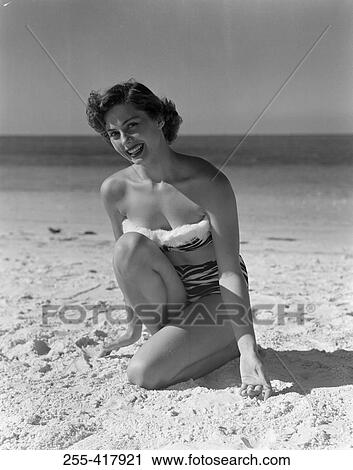 Stock Image - Portrait of pin-up girl wearing bikini, crouching on sandy beach. Fotosearch
