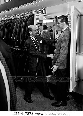 Stock Image - Shop assistant serving man trying on suit in shop. Fotosearch