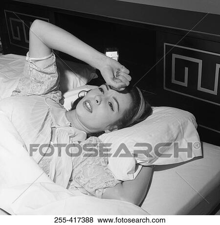 Stock Photo - Young woman lying in bed with hand on forehead. Fotosearch