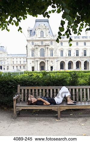 Stock Photography - Young woman resting in a garden with a museum in the background, Jardin De Tuileries, Musee Du Louvre, Paris, Ile-De-France, France. Fotosearch