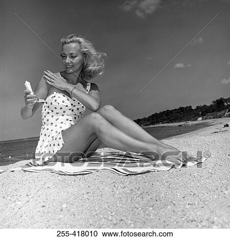 Stock Image - Young woman sitting on beach and applying suntan lotion. Fotosearch