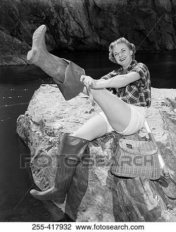 Stock Image - Young woman sitting on rock putting on wellington boots. Fotosearch