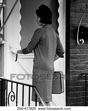 Stock Image - Young woman standing in front of entrance door using doorbell. Fotosearch