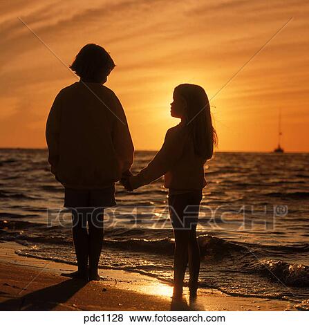 Silhouette Of A Young Boy And Girl Holding Hands On The Shore As The Sun Sets Into The Ocean In Front Of Them Stock Photo Pdc1128 Fotosearch