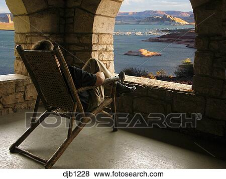 Man Relaxing On Lounge Chair Seen From Behind In Front Of Opening In Rock Porch With Scenic Lake Boats And Mountains In Background Lake Powell