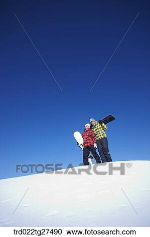 Stock Image - couple standing on the snow hill with a snow board. Fotosearch
