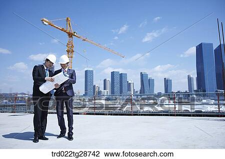 Stock Image - busineess people are going throught the blueprints at construction site. Fotosearch