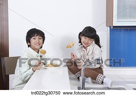 Stock Image - Happy son and daughter eating bread in the kitchen. Fotosearch