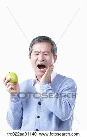 Stock Image - A painful elderly man with toothache while holding an apple . Fotosearch