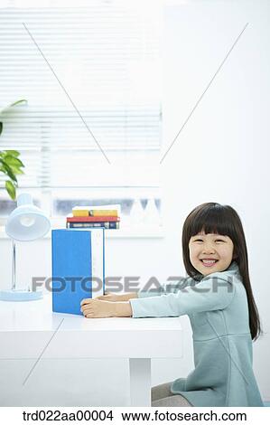 Picture - A smiling girl having a book in front of the desk. Fotosearch