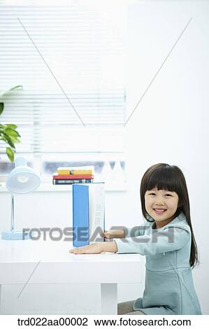 Stock Image - A smiling girl having a book in front of the desk. Fotosearch