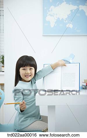 Stock Photograph - A smiling girl showing a book in front of the desk. Fotosearch
