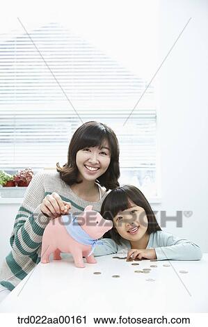 Stock Image - A woman and girl putting a coin into a pink piggy bank. Fotosearch