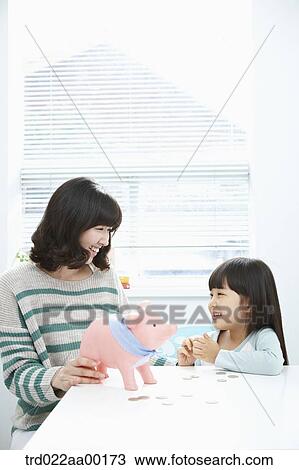 Stock Image - A woman and girl smiling at each other with a pink piggy bank and coin on the desk. Fotosearch