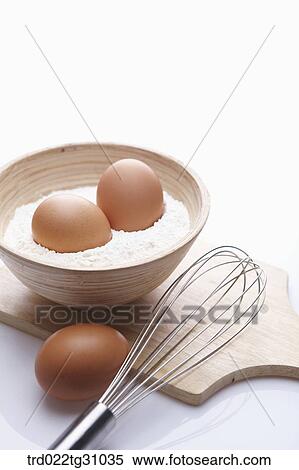 Stock Photography - The eggs in the bowl of flour and the whisk on the cutting board. Fotosearch
