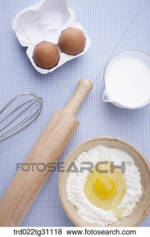 Stock Photo - The eggs in the carton, whisk, rolling pin, a cup of milk, and egg in the bowl of flour. Fotosearch