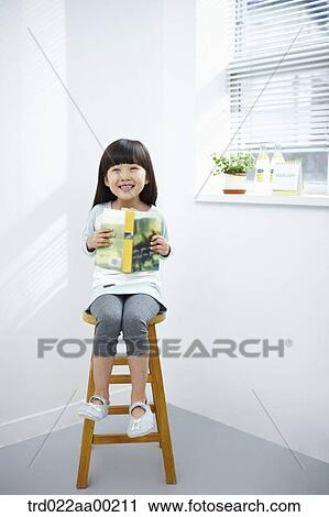Stock Image - The girl holding a book on the chair. Fotosearch