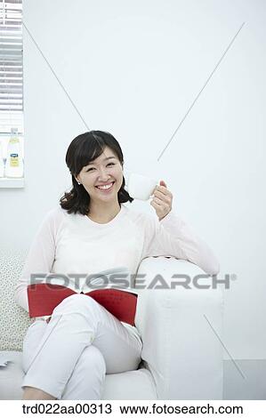 Stock Image - The woman holding the cup of coffee and book on the couch. Fotosearch