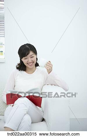 Stock Photography - The woman holding the cup of coffee and book on the couch. Fotosearch