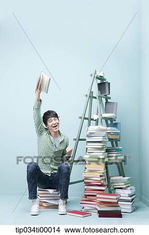 Picture - A man holding up a book by the piled books and ladder. Fotosearch