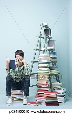 Stock Image - A man reading a book by the piled books and ladder. Fotosearch