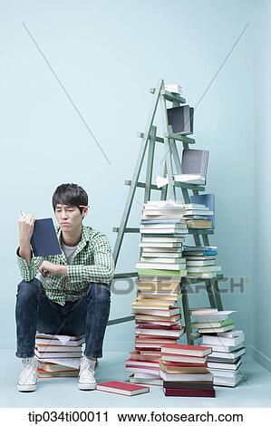 Stock Image - A man reading a book by the piled books and ladder. Fotosearch