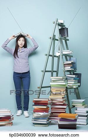 Stock Photo - A woman holding up a book by the piled books and ladder. Fotosearch