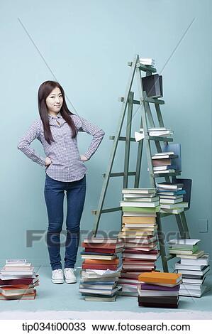 Stock Image - A woman standing by the piled books and ladder. Fotosearch