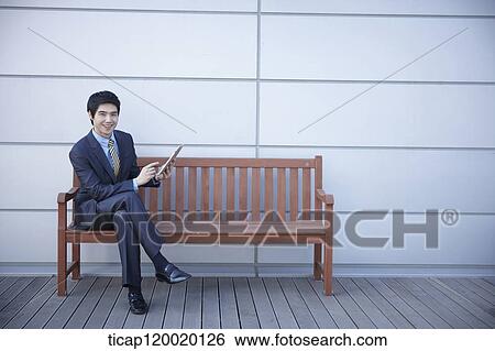 A businessman sitting on a bench holding a touch pad View Large Photo Image Stock Photograph - A businessman sitting on a bench holding a touch pad. Fotosearch
