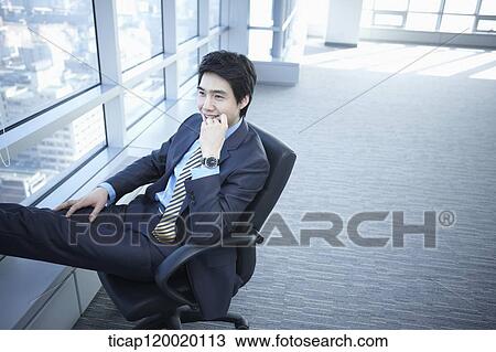 Stock Image - A businessman sitting on a chair leaning his lengs on window. Fotosearch