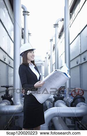 Stock Image - A female architect smiling holding a blue print next to pipeline. Fotosearch
