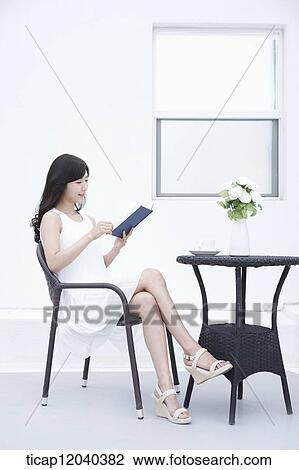 Stock Image - A woman in a white dress sitting on black garden furniture and reading a book. Fotosearch