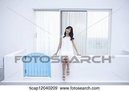 Stock Photo - A woman in white dress sitting on a white fence smiling. Fotosearch