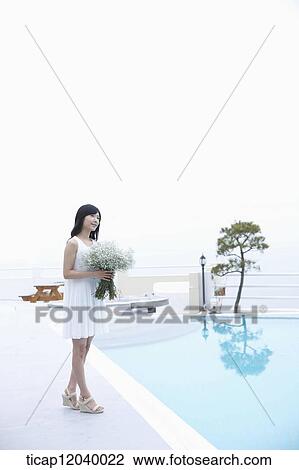 Stock Image - A woman in white dress standing with a flower bouquet next to a pool. Fotosearch