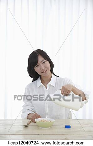 A woman in white shirt sitting at a dining table pouring milk to a bowl of cereal View Large Photo Image Picture - A woman in white shirt sitting at a dining table pouring milk to a bowl of cereal. Fotosearch