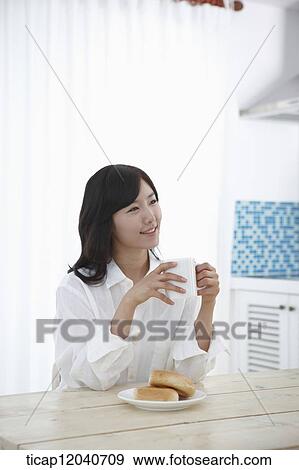 Stock Photo - A woman in white shirt sitting at a table drinking a cup of coffee . Fotosearch