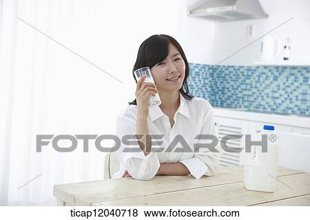 Stock Photo - A woman in white shirt sitting at a dining table holding a cup of milk next to her face. Fotosearch