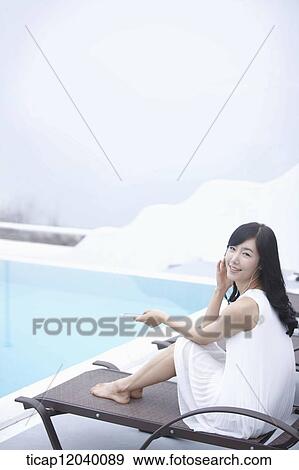 Stock Photo - A woman sitting on a sunbed next to a pool smiling. Fotosearch
