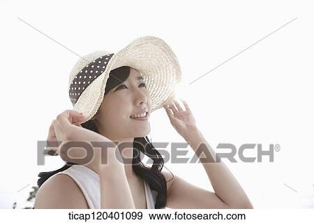 Stock Photo - A woman wearing a white dress and straw hat grabbing her hat with both hands. Fotosearch