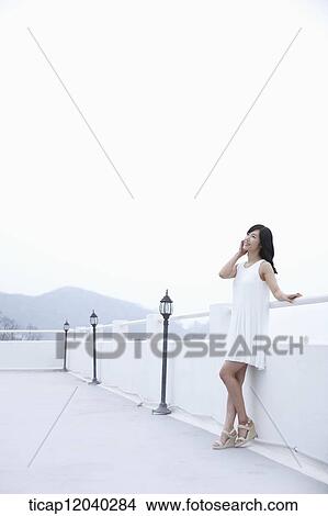 Picture - A woman with a white dress leaning on a white fence next to a black garden light. Fotosearch