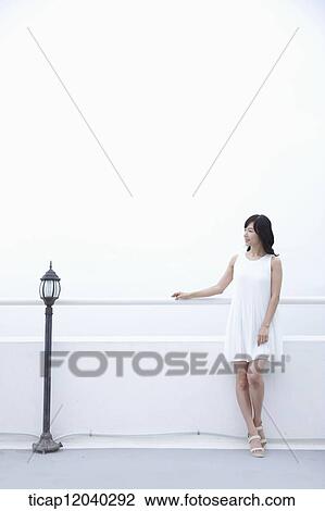 Stock Image - A woman with a white dress leaning on a white fence next to a black garden light. Fotosearch