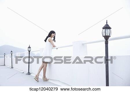 Stock Photo - A woman with a white dress leaning on a white fence and watching the view. Fotosearch