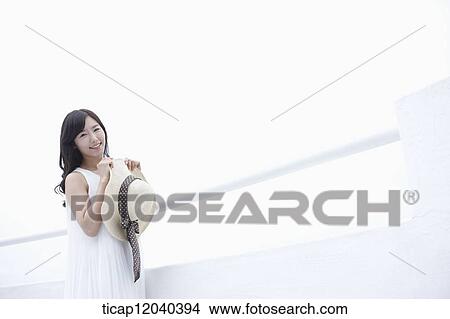 A woman with a white dress standing next to a white fance with a sun hat in her hands while looking towards the camera View Large Photo Image Picture - A woman with a white dress standing next to a white fance with a sun hat in her hands while looking towards the camera. Fotosearch