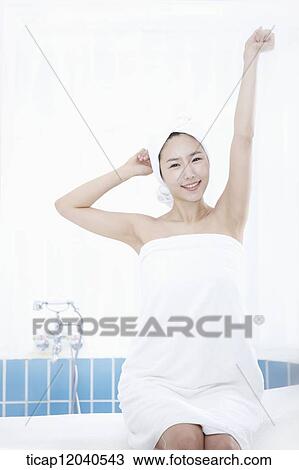 Stock Image - A woman with bath towel around body and over head sitting at edge of bath tube. Fotosearch