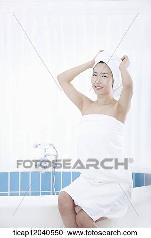 Stock Image - A woman with bath towel around body and over head sitting at edge of bath tube. Fotosearch
