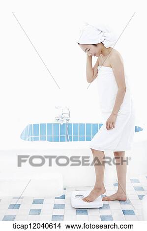 Picture - A woman with bath towel around her body and over head weighing herself on a scale. Fotosearch