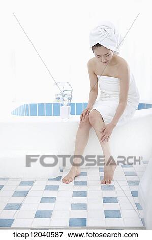 Stock Photo - A woman with bath towel around her body and over head sitting at edge of bath tub putting lotion on her leg. Fotosearch