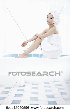 Stock Photograph - A woman with bath towel around her body sitting with two feet on a edge of a bathtub. Fotosearch
