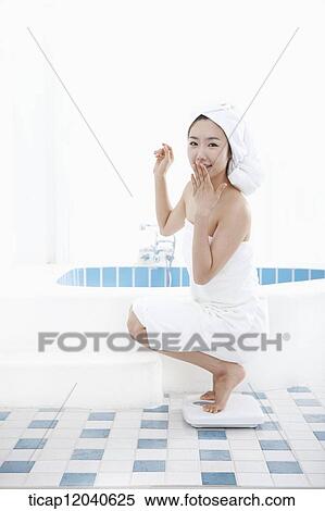Stock Photography - A woman with bath towel around her body and over head weighing herself with two feet on a scale. Fotosearch